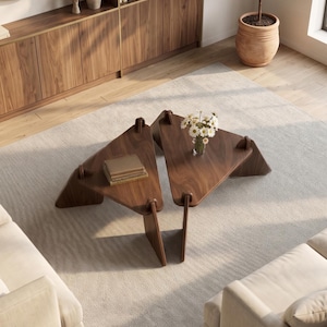 May include: Two triangular wooden coffee tables with a dark finish are arranged on a light gray rug. One table holds a stack of books, the other a vase of white flowers. The room features a wooden cabinet, a mirror, and a potted plant.