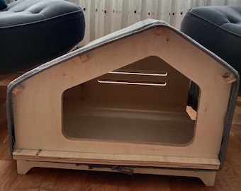 May include: A wooden pet house with a gray fabric roof. The house has a rectangular opening and sits on small wooden legs. The interior is visible through the opening. The house is designed to resemble a small house.