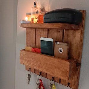 May include: A wooden wall-mounted organizer with a shelf, a pocket, and key hooks. The shelf holds a black glasses case, candles, and perfume bottles. The pocket contains a phone and keys. Keys hang from the hooks.