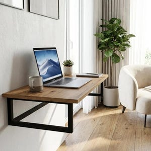 May include: A wall-mounted floating desk featuring a rectangular wooden top with a natural grain finish and a black metal frame. The desk holds a silver laptop, a two-toned ceramic mug, a small potted succulent, and a flat notebook on its surface.