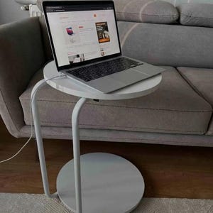 May include: A white metal side table with a circular top and a matching circular base, featuring a C-shaped frame. A silver laptop is placed on the top surface of the table, with a white charging cable connected to the side of the laptop.
