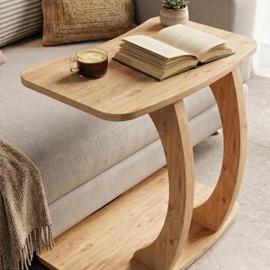 May include: A light wood C-shaped side table with a rectangular top and a curved base support. The table surface holds an open book, a glass mug filled with dark coffee, and a small potted succulent plant, showcasing a functional furniture piece.