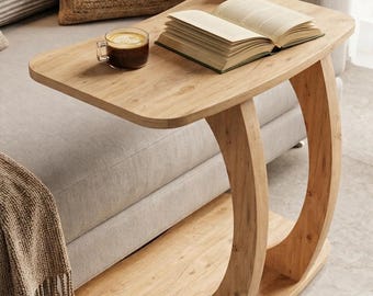 May include: A light brown wooden C-shaped side table with a smooth rectangular top and a curved base support. An open book, a glass mug filled with dark coffee, and a small potted succulent plant are placed on the surface of the wooden table.