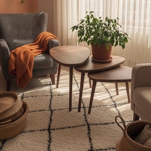 May include: A cozy living room scene featuring a gray armchair with an orange throw blanket, a set of three wooden side tables, and a patterned rug. A potted plant sits atop the tables. Woven baskets add to the warm, inviting atmosphere.