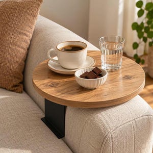 May include: A round wooden sofa arm table with a natural wood grain finish and a black metal support bracket. The table surface holds a white ceramic coffee cup on a matching saucer, a clear glass of water, and a small white bowl containing chocolate.