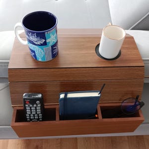 Puede incluir: Una mesa de reposabrazos de sofá de madera con portavasos y compartimentos de almacenamiento integrados. Contiene una taza azul y blanca de temática náutica, una taza blanca, un mando a distancia, un cuaderno azul y gafas. La mesa es marrón con detalles dorados.
