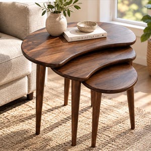May include: A set of three nesting tables made of dark brown wood with a smooth finish. Each table features a kidney-shaped top and three tapered legs. A beige ceramic vase, a small speckled bowl, and a book titled The Kinfolk Home sit on top.
