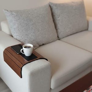 Könnte beinhalten: Ein dunkles Sofa-Armtablett aus Holz mit schwarzem Einsatz hält eine weiße Kaffeetasse und ein Smartphone. Das Tablett steht auf der Armlehne eines weißen Sofas, im Hintergrund zwei grau gemusterte Kissen.