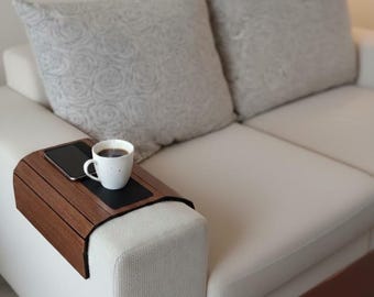 Könnte beinhalten: Eine flexible Armlehnenablage aus dunkelbraunem Holz mit einem rechteckigen schwarzen Inlay in der Mitte. Die Ablage liegt auf der Armlehne eines hellbeigen Sofas und trägt eine weiße Keramiktasse mit schwarzem Kaffee sowie ein schwarzes Smartphone.