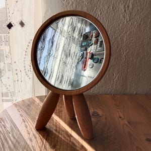 Può includere: Uno specchio da toeletta rotondo in legno con una base a treppiede. Lo specchio ha una cornice in legno naturale e una superficie riflettente. Lo specchio è posto su un tavolo di legno. Lo sfondo include una tenda trasparente e un muro.