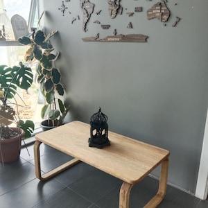 Op de afbeelding: Een rechthoekige salontafel van licht hout met afgeronde randen en doorlopende luspoten. Een zwarte metalen lantaarn met een ingewikkeld uitgesneden patroon en een koepelvormige bovenkant staat in het midden van het tafeloppervlak voor kleurcontrast.