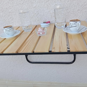 May include: A small wooden table with a black metal frame, holding two cups of coffee with saucers, two glasses of water, a small dish, and a glass bowl with pink candies. The table is set against a white wall.