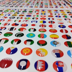 May include: A sheet of numerous small, circular, glossy stickers featuring various national flags from around the world. The stickers are arranged in a grid pattern on a white backing, each displaying distinct colors and symbols representing different countries.