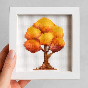 May include: A framed cross-stitch artwork featuring a tree with a full, vibrant yellow and orange canopy. The tree's trunk and branches are brown, set against a white background. The artwork is encased in a square white frame.