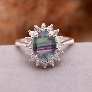 Può includere: Anello in argento con una pietra di fluorite ovale multicolore. La pietra presenta sfumature di blu, verde e viola. È circondata da un'aureola di piccole pietre chiare. Un anello elegante e accattivante.