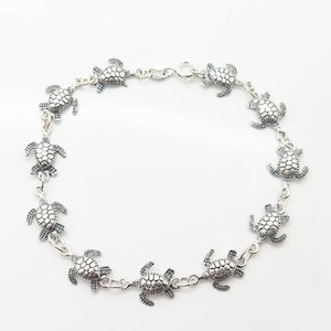 Può includere: Bracciale argentato con una serie di piccoli ciondoli a forma di tartaruga. Ogni tartaruga presenta dettagli sul guscio e sulle pinne, collegati da delicati anelli. Il bracciale ha una chiusura a moschettone.