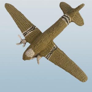 May include: A knitted olive green airplane toy with white and black striped accents on the wings and tail. The propeller is silver. The toy is made of soft yarn and is suitable for children.