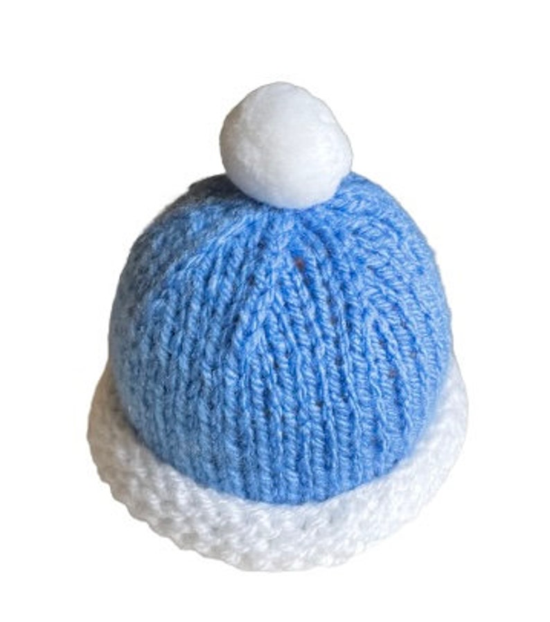 May include: A blue knitted baby hat with a white pom pom on top. The hat has a white brim.