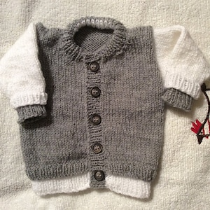 Puede incluir: Un cárdigan de punto gris con mangas blancas y botones marrones. El cárdigan está colocado sobre una superficie blanca.