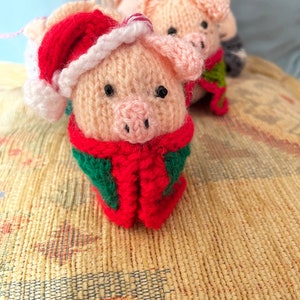 May include: Two knitted pig ornaments, one wearing a red Santa hat and a green scarf, the other wearing a green scarf. Both are on a beige textured surface.