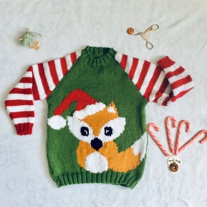 May include: A green knitted sweater with a fox wearing a Santa hat. The sweater has red and white striped sleeves. Candy canes and other holiday decorations are arranged around the sweater.