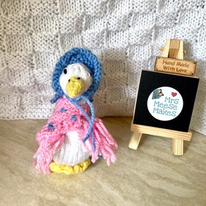 May include: A knitted goose wearing a blue bonnet and a pink scarf. The goose is handmade and has a sign that says "Hand Made With Love" and "Mrs MeeSe Makes".