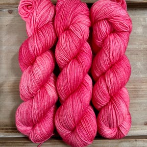 Könnte beinhalten: Drei Stränge leuchtend rosa Garn, fest zu verdrehten Bündeln gewickelt. Das Garn hat ein weiches, strukturiertes Aussehen mit subtilen Farbvariationen. Der Hintergrund ist eine Holzoberfläche. Ideal für Strick- und Häkelprojekte.