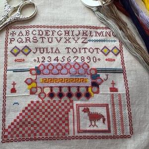 May include: A cross-stitch sampler with the name "JULIA TOITOT" and numbers. The sampler features a red and white checkered pattern, a framed animal, and a decorative border. Silver scissors and colorful embroidery floss are also present.