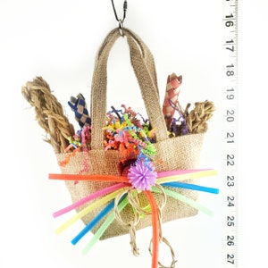 May include: A small, burlap basket filled with colorful paper shreds, rope, and woven items. The basket has a handle and is decorated with rainbow-colored straws and a purple pom-pom. The basket is suspended from a black cord.