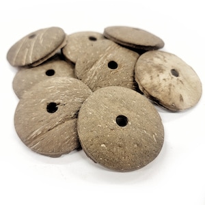 May include: A collection of natural, round coconut shell beads. Each bead is a flat disc shape with a central hole, and a textured, brown surface. The beads are scattered on a white background, showcasing their organic texture and color.