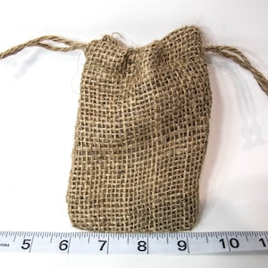 May include: A small, rectangular burlap drawstring bag. The bag is a natural tan color with a woven texture. The drawstring is made of twisted twine. The bag is closed at the top with the drawstring pulled tight.