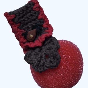 May include: A handmade, red and black crocheted kitchen sponge with a decorative holder. The holder features a star-shaped button and a bow. The sponge is round and has a textured appearance, with a touch of sparkle.