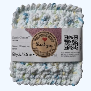 Könnte beinhalten: Ein quadratisches Knäuel aus weißem, gehäkeltem Baumwollgarn mit blauen, gelben und grünen Flecken. Das Etikett trägt die Aufschrift "Classic Cotton" und ein Aufkleber "thank you". Das Garn ist 71 g.