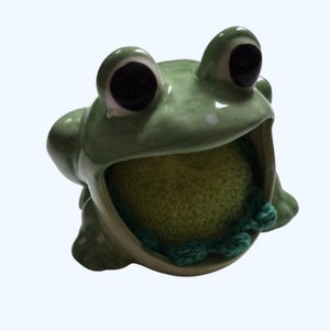 May include: A ceramic frog-shaped sponge holder in a light green glaze. The frog has large black eyes and a wide open mouth holding a green sponge. The holder is designed to sit on a countertop.