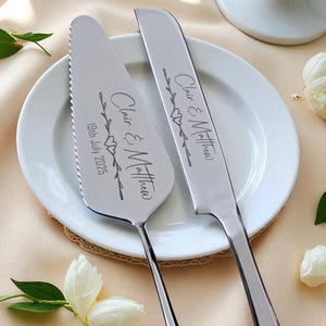 Może przedstawiać: Srebrny zestaw do serwowania ciasta i noża na białym talerzu. Serwer ma ząbkowaną krawędź, a zestaw jest wygrawerowany napisem "Clair & Matthew" i datą "19 lipca 2025". Dekoracyjne akcenty kwiatowe są zawarte w grawerunku. Zestaw jest na wesele.