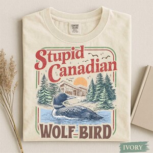 以下が含まれることがあります： 「Stupid Canadian Wolf-Bird」の文字が入ったグラフィックが特徴のアイボリー色のTシャツ。水面にいるハシグロアビ、小屋、松の木、夕焼けのデザイン。Tシャツは折りたたまれています。