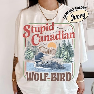 以下が含まれることがあります： アイボリー色のTシャツ。水面にいるハシビロガモ、松の木、そして「Stupid Canadian Wolf Bird」の文字が描かれたグラフィック。デザインには夕日と建物が含まれています。シャツには「Comfort Colors」と「Ivory」がプリントされています。