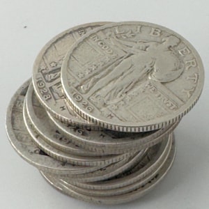 May include: A stack of silver coins, each displaying a detailed design with the word "LIBERTY" and the year 1926 or 1928. The coins are arranged in a slightly tilted pile, showcasing their edges and intricate details.