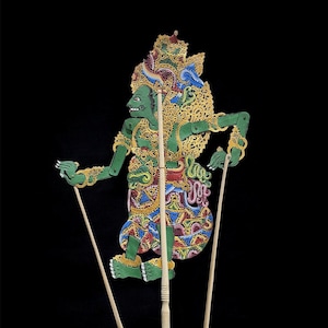 Può includere: Un burattino delle ombre balinese verde e oro con dettagli intricati. Il burattino ha un copricapo dettagliato e tiene in mano delle bacchette di controllo in legno. Il burattino è su una base di legno su sfondo nero.