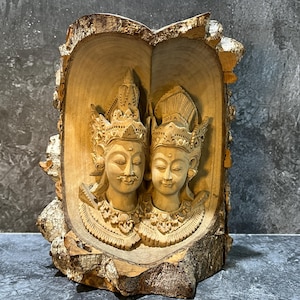 以下が含まれることがあります： 装飾的なヘッドドレスと詳細なジュエリーを身に着けた、穏やかな顔が2つ描かれた木彫りの彫刻。彫刻は天然木のフレーム内にあり、樹皮の質感と滑らかに彫られた姿が特徴です。