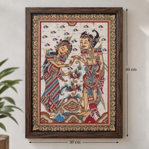 Op de afbeelding: Een ingelijst schilderij met twee figuren in traditionele kledij te midden van gestileerde florale elementen. Het kunstwerk zit in een donkerbruine houten lijst van 30 cm bij 40 cm, met ingewikkelde patronen in rode, blauwe en gouden tinten.
