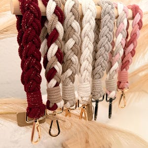 Könnte beinhalten: Eine Sammlung geflochtener Schlüsselbänder in verschiedenen Farben, darunter Dunkelrot mit goldenem Metallic-Faden, Beige, Creme, Grau und Rosa. Jedes Band hat ein dickes, gewebtes Kordeldesign und ist an einem Karabinerhaken aus Metall befestigt.