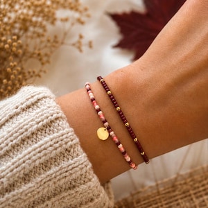 Könnte beinhalten: Zwei zarte Perlenarmbänder. Ein Armband besteht aus einem Farbverlauf aus rosa, roten und weißen Perlen mit einem kleinen, runden, goldfarbenen Metallanhänger. Das zweite Armband besteht aus dunkelburgunderroten Perlen mit goldenen Akzenten.