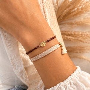 Könnte beinhalten: Zwei Armbänder, die zusammen getragen werden. Das obere Armband ist eine dünne dunkelbraune Kordel mit einem kleinen goldfarbenen Ammoniten-Anhänger. Das untere Armband ist ein breiteres beiges Webband mit Chevron-Muster und einer Quaste.
