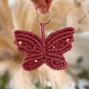 Könnte beinhalten: Ein handgefertigter, staubrosa Makramee-Schmetterling-Schlüsselanhänger. Der Schmetterling hat detaillierte Flügel mit kleinen Öffnungen und ist an einem silbernen Metall-Schlüsselring befestigt. Der Schlüsselanhänger wird vor einem verschwommenen Hintergrund aus getrockneten Pflanzen gehalten.