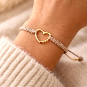 Könnte beinhalten: Ein Armband aus beigefarbenem geflochtenem Kordelmaterial mit einem mittigen, polierten, goldfarbenen Herzanhänger. Das Armband verfügt über einen verstellbaren Schiebeknotenverschluss mit zwei herabhängenden Kordelenden für einen sicheren Sitz.