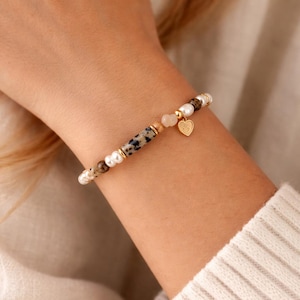 Könnte beinhalten: Ein Armband mit Perlen, gesprenkelten Perlen und goldenen Akzenten. Ein goldener Herzanhänger hängt an dem Armband. Das Armband wird an einem Handgelenk vor einem neutralen Hintergrund getragen.