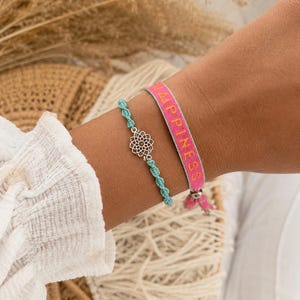 Könnte beinhalten: Zwei Armbänder zusammen getragen. Eines ist ein hellblaues geknotetes Kordelarmband mit einem silberfarbenen Blumenanhänger aus Metall. Das zweite ist ein flaches, gewebtes rosa Armband mit dem Wort HAPPINESS in gelbem Garn und einer silberfarbenen Metallperle.