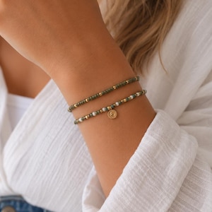 Könnte beinhalten: Zwei Armbänder mit Perlen. Das obere Armband hat olivgrüne und goldene Perlen. Das untere Armband hat weiße, olivgrüne und goldene Perlen mit einem kleinen goldenen Sonnenanhänger. Die Armbänder werden am Handgelenk getragen.