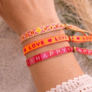 Könnte beinhalten: Drei gewebte Stoffarmbänder mit goldenen Metallic-Faden-Akzenten. Das obere Band zeigt eine gelbe Sonne und geometrische Muster. Das mittlere orangefarbene Band trägt den Text LOVE mit Herzsymbolen. Das untere rosa Band zeigt das Wort HAPPY.